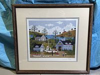 Photo shows framed signed and numbered print with cream mat and dark wood frame. The image is a coastal village with houses, lighthouse, and people.
