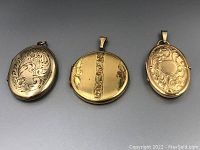 Three gold-colored vintage lockets displayed side by side, showing front engraved designs and bails.
