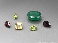 Six loose gemstones displayed on gray background including garnets, peridots, citrine, and green cabochon stone