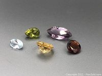 Five loose faceted gemstones arranged on a grey background showing size, cut and color variety.