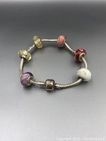 Image showing full round silver-tone bracelet with six assorted glass bead charms featuring various colors and designs on neutral gray background.