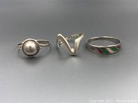 Photo showing three sterling silver rings laid flat against a neutral background. The first ring has a round domed center, the middle ring is a V-shaped design, and the third ring features enamel inlay with red, green, and white colors.