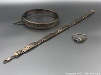 Three sterling silver jewelry pieces on gray background: hinged bangle bracelet with engraved swirls and safety chain, geometric patterned flat link bracelet, and ring with central clear stone and side accents.