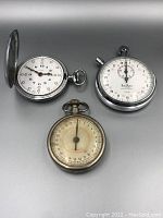 Three vintage timepieces: one Cyma pocket watch with cover and two Hanhart stopwatches, all showing signs of age and wear.