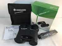 Vanguard Orros 8320 binoculars with carrying case, user manual, neck strap in plastic, and original box displayed on white surface.