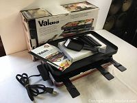 Full view of the SWISSMAR Valais Red Raclette grill with electrical cord, 8 raclette pans and instruction manual next to original box.