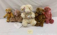 Full line up of five collectible stuffed plush animals against a white backdrop, centered in frame. From left to right: beige bear, curly brown bear with a bow, white bunny with basket, brown teddy bear with Ty tag, and pink teddy bear with a pink ribbon.