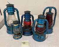 Five vintage kerosene barn lanterns with blue metal bodies, including red and clear glass globes, arranged on table.