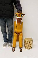 Front view of the tall seated Native American wooden sculpture with painted headdress and small drum beside it for scale.