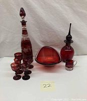 Overview of the entire glassware set showing all items including the tall Amberina bottle with stopper, ruby red Murano dish, Bohemian decanter, and six cordial glasses with etched floral designs.