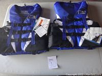 Two new Fluid brand adult small life jackets side by side with tags attached, showing front view of jackets, highlighting three adjustable black strap buckles and blue color.