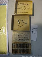 Photo of a yellow Milrone Enameled Compacts box open, containing five small metal watch stems or parts, and a wooden box with multiple compartments beneath it.