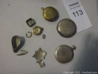 Photo showing multiple pocket watch cases and small scrap pieces on a white surface with a lot number card