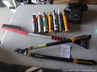Full lot with black bakelite rotary dial phone, red and black brush, seven tubes of caulking, and Fiskars tree limb pruner on a grey table