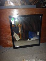 Frontal view of the black wood framed mirror leaning against a wooden backdrop, showing the reflective glass surface of the mirror.