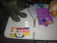 Wide view of green rubber waders, fishing tackle box, and net on white table