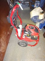 Side view of red and black Briggs & Stratton gas pressure washer on wheels with attached spray wand and detergent tank.