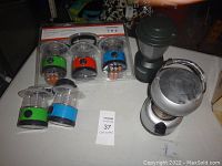 Photo showing three new small battery operated lanterns in package (orange, green, blue) and five larger used battery operated lanterns of various colors and styles including a black Coleman lantern.