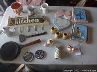 Full view of assorted kitchen and decorative items on a table including ceramic planter, mugs, cast iron sign, figurines, and plaques.