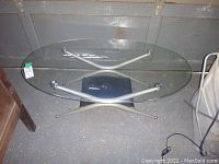 Oval glass top coffee table with silver metal crossed-leg frame on a concrete floor, shown from an angle.