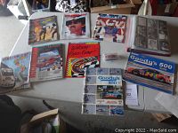 Wide shot of eleven programs, magazines, and a diecast car displayed on a tabletop, showing covers with racing images and car models.