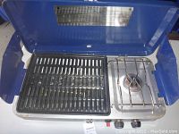 Open view of the blue propane camping stove and grill showing the single burner and grill section with metal grates and side wind guards.