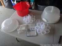 Photo of all items on table including red cake carrier base, three clear stacking cupcake trays, clear dome lid, small plastic cups, instruction booklet, and small packet.