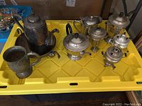 Photo of nine metal teapots, samovars, and a cup arranged on a yellow plastic surface