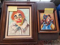 Photo showing two clown paintings framed, larger one has orange hair and white ruffled collar, smaller has jester hat and blue outfit.