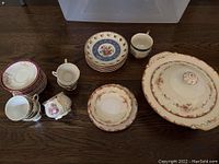 Overview of various plates, tea cups, saucers, and a large covered serving dish, arranged on a wooden surface.