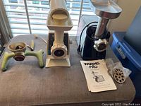 Photo of all three meat grinders on a table. Left is the antique hand grinder with green cast iron body, center is the cream-colored mid-century Oster electric grinder, right is the modern black Waring Pro grinder with additional discs and instruction manual.
