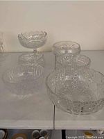 Photo showing six clear crystal serving bowls arranged on a table with visible detailed cut glass patterns and varied shapes including a tall pedestal bowl and several round and oval bowls.