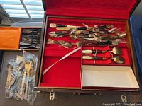 Wide view of large wooden briefcase opened showing assorted antique silverware, with some pieces also packaged in plastic on table.