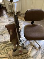 Photo showing the Royal Heavy Duty vacuum cleaner with its brown cloth bag and metal body, standing next to the brown upholstered desk chair on caster wheels.