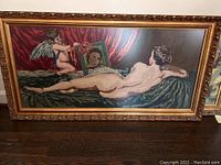 Full view of the large crochet knit painting framed in gold showing the reclining woman, cherub, mirror, and draped fabric.