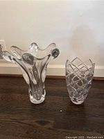 Two clear glass vases on dark wood floor. Left vase is tall with wavy top, right is Mikasa vase with geometric cuts.