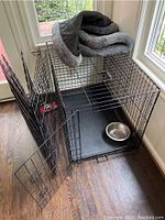 Black metal wire dog cage with open door, showing removable plastic base tray inside and a stainless steel bowl for size reference; grey dog bedding is visible on top of cage but not included in lot.