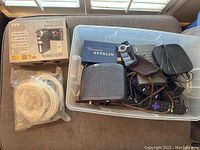 Photo of varied electronics including a Netgear black device, video cameras, cables, and remote controls in bin