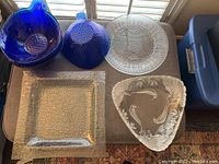 All items displayed showing the three serving trays: one clear round plate with crackle texture, one clear triangular plate with raised fish design, and one square amber textured plate. Also displayed are five deep blue glass bowls with embossed fish and sea life designs.
