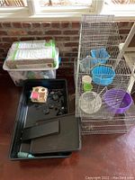 Photo showing two large metal wire hamster cages with black plastic bases. One cage contains blue bowl, purple exercise wheel, clear water bottle, and small platforms. A plastic bin with bedding and supplies and a hamster house also visible.