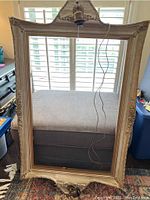 Full view of ornate wooden lighted picture frame leaning against window with blinds. Hanging cord and bulb socket visible at top center.