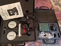 Fluorescent leak detection kit with two analog gauges, hoses, valve assembly, and instruction booklet in a molded plastic case.