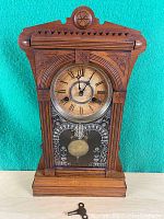 Full frontal view of the antique gingerbread clock with wooden carved case, glass front showing pendulum, and winding key placed beneath on light wood surface.