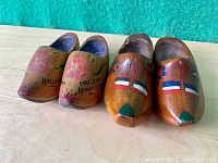 Two pairs of painted miniature wooden Dutch clogs shown side by side on a tan surface with green background.
