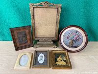 Five antique frames arranged on table; one with needlework floral picture; others hold vintage black and white photos