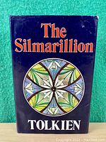 Front cover of The Silmarillion book with circular stained glass floral design and title in orange and white text on dark blue background.