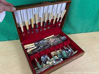 Wooden chest with various flatware and serving pieces arranged inside, including ivory-handled knives in the lid and multiple spoons and forks in the base compartment.