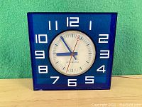 Front view of the cobalt blue square plastic framed General Electric kitchen clock with white face and blue hour and minute hands, red second hand.