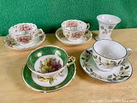 Four vintage floral teacups with matching saucers and one Wedgwood vase with blue floral detailing on wood surface with green background.