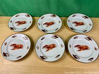 Six vintage porcelain plates with lobster motifs and seashells surrounding them, arranged in two rows of three on a wooden surface with green background.